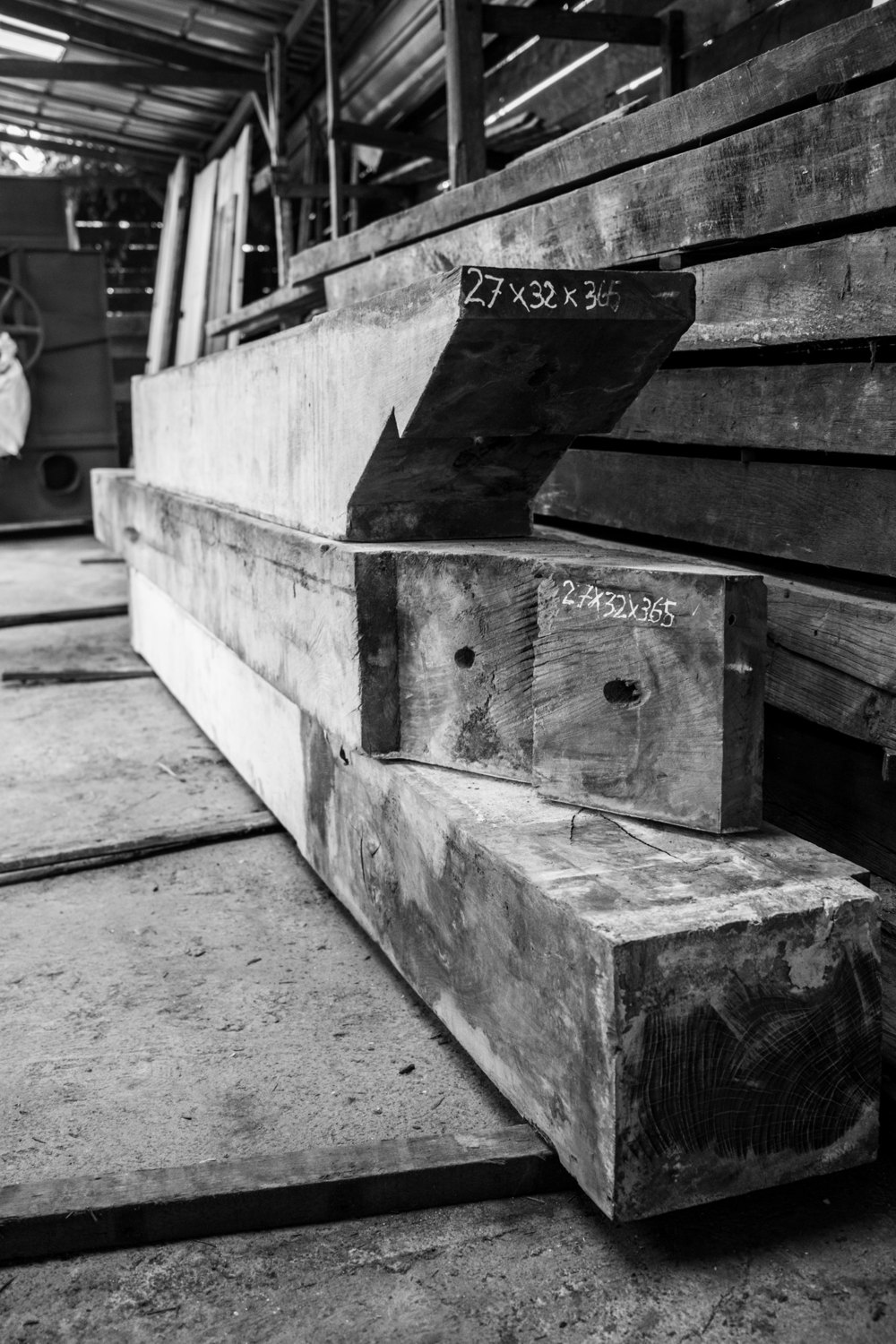 Black-and-white photograph of heavy old bridge beams stacked in a timber shed, their sizes written in chalk on the cut ends