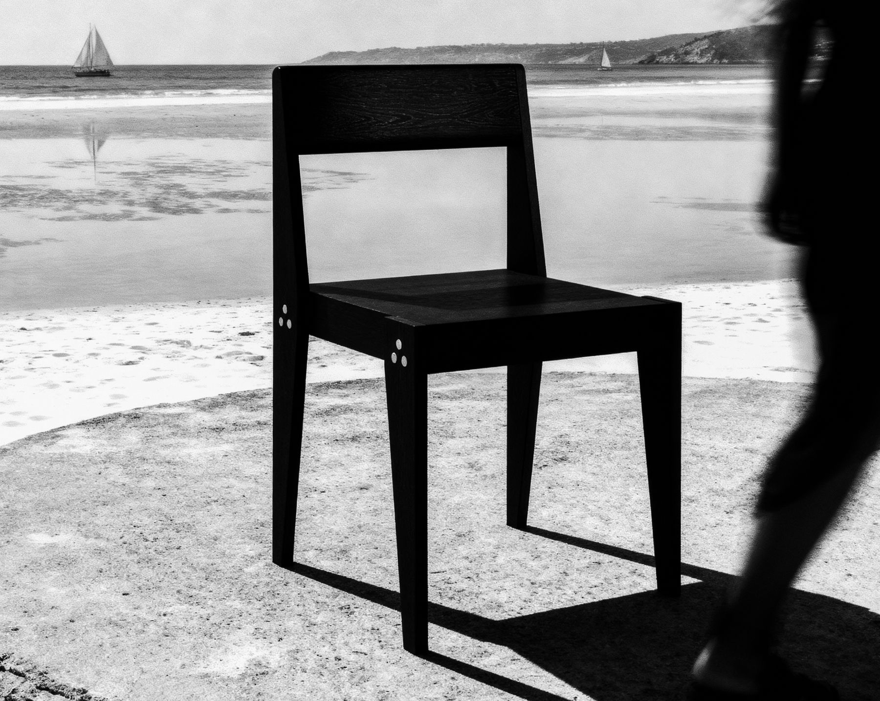 Black-and-white photograph of The Chair on a sea wall at low tide, a sailboat on the water behind and a figure passing out of focus