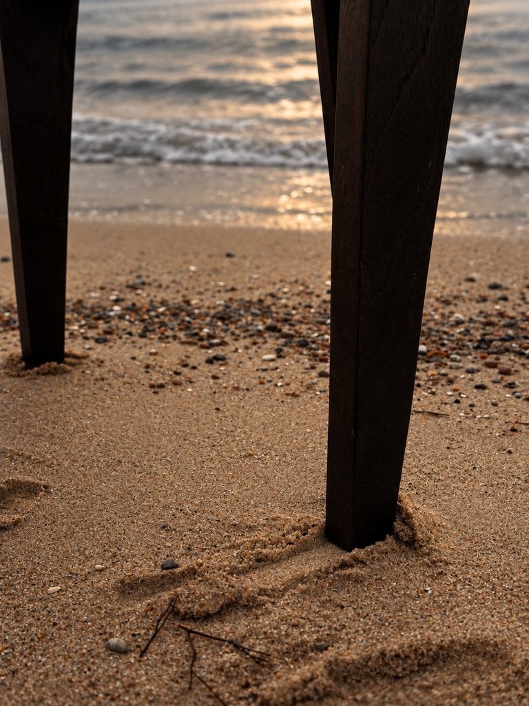 Close-up of two dark teak chair legs standing in sand at the water's edge, small waves behind