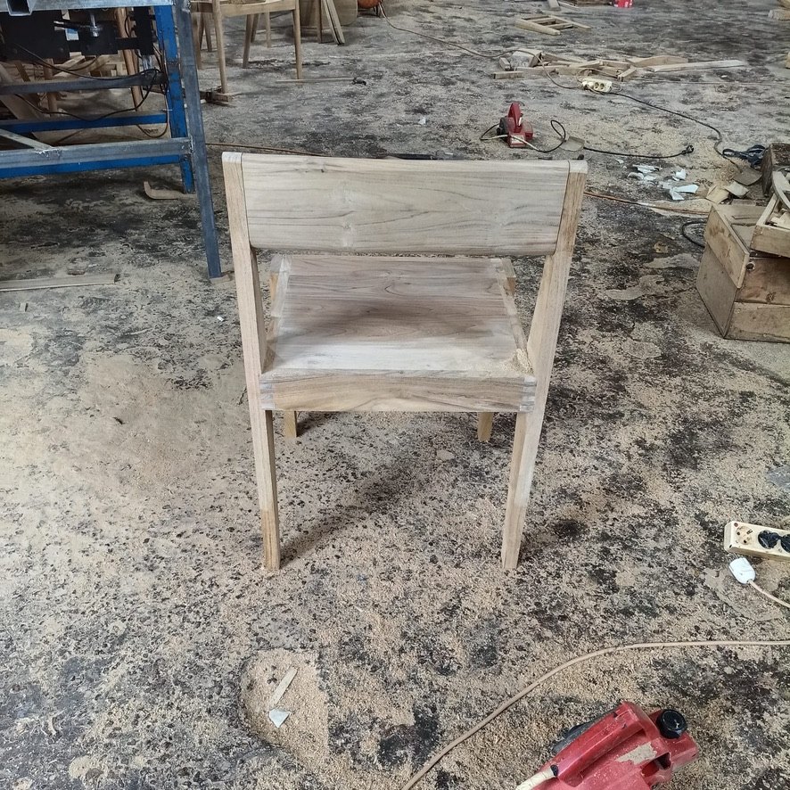An unfinished teak chair standing on a sawdust-covered workshop floor, before burning