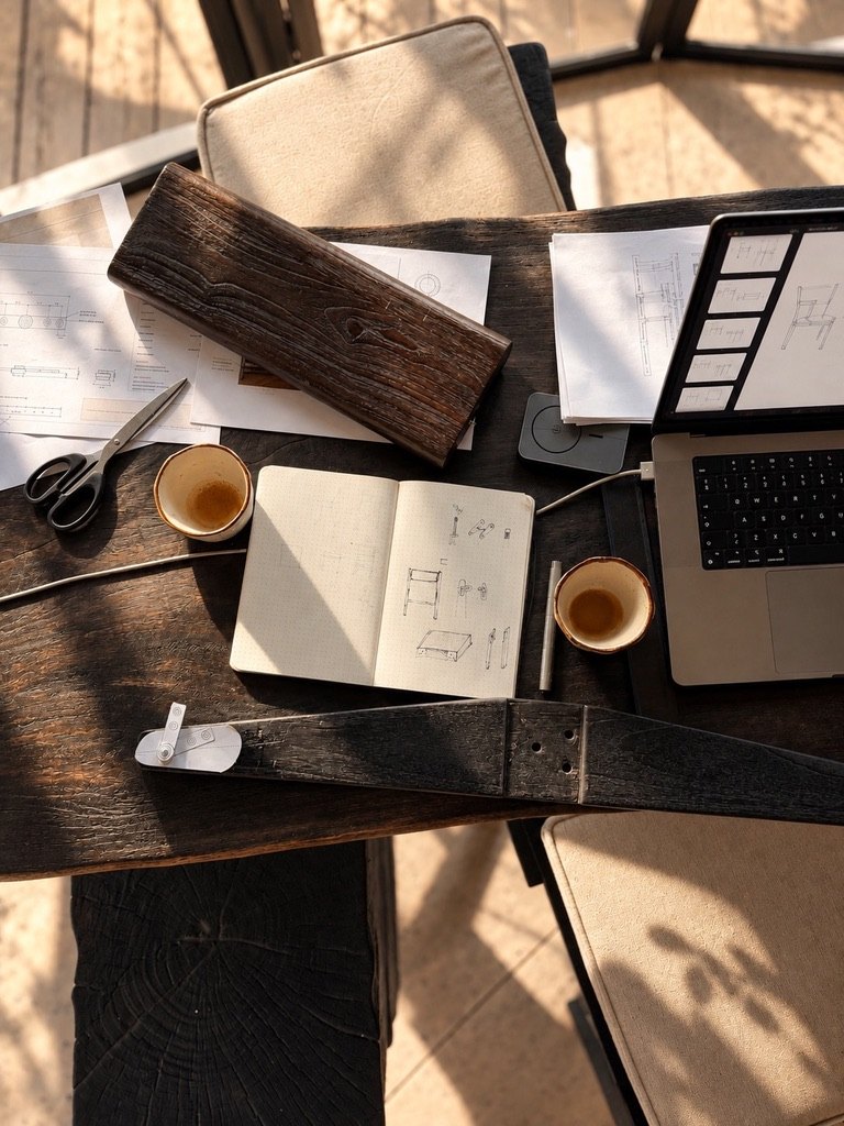 A dark timber table in afternoon sun with chair drawings, a sketchbook, a laptop showing chair designs, a burned teak sample and two coffee cups