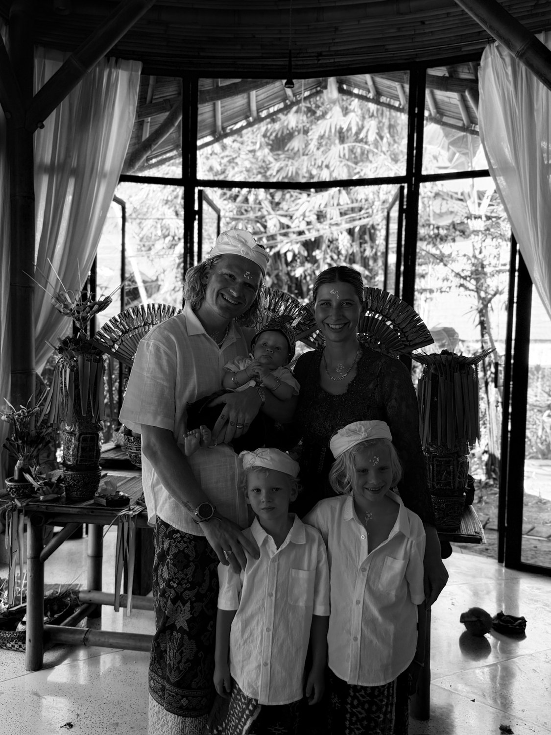 Black-and-white photograph of the founders and their three children in Balinese ceremonial dress, smiling, with offerings and a garden behind them
