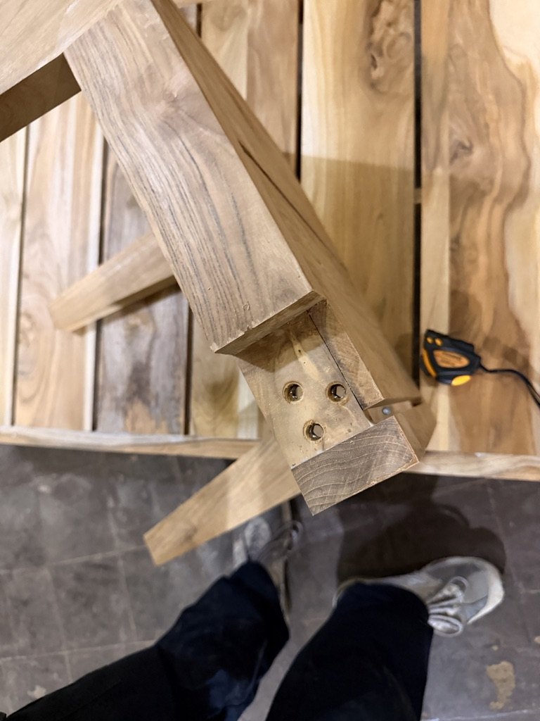 The underside of a raw teak frame, showing a leg joint with three bolt holes and the threaded inserts set into the wood