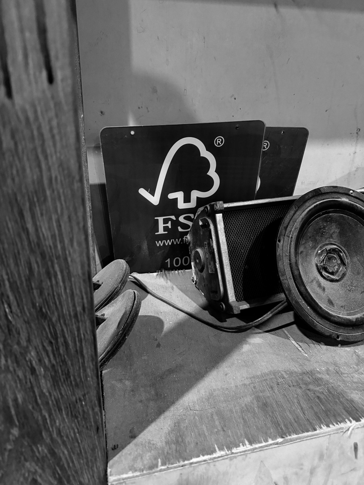 Black-and-white photograph of certification signs leaning against a workshop wall, next to an old speaker and a pair of work gloves
